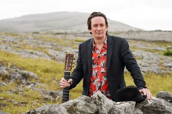 A blind musician holds a guitar in rolling Irish hillside